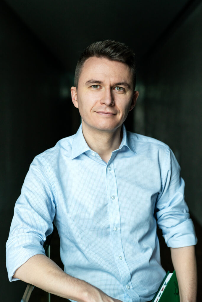 Uśmiechnięty mężczyzna stojący frontem do aparatu z zielonym notesem w ręku